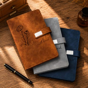 May include: Three rectangular notebooks in brown, gray, and navy blue with a matte finish. Each notebook features a magnetic strap with a silver rectangular accent, a laser-engraved floral illustration, and the name David in cursive script.