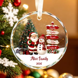 May include: A round, clear glass Christmas ornament featuring a printed scene of Santa Claus and a snowman standing next to a decorated tree. A signpost displays the names Lucas, Lydia, Oscar, Ella, and Cookie. Text reads Alice family 2026.