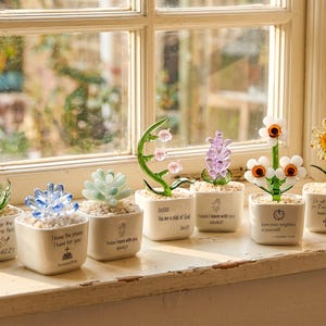 May include: A collection of six small glass plants in white square ceramic pots, each featuring a unique flower or succulent design and a printed religious verse. The plants include green and blue succulents, a lily of the valley, and daisies.