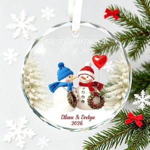 May include: A round, clear glass ornament featuring a printed graphic of two snowmen wearing blue and red winter hats and scarves. The text reads FIRST CHRISTMAS AS Mr. & Mrs., Ethan & Evelyn, and 2026. A red heart balloon is held by one snowman.