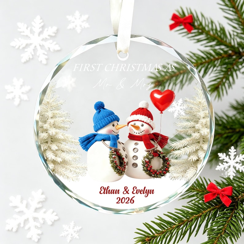 May include: A round, clear glass ornament featuring a printed graphic of two snowmen wearing blue and red winter hats and scarves. The text reads FIRST CHRISTMAS AS Mr. & Mrs., Ethan & Evelyn, and 2026. A red heart balloon is held by one snowman.