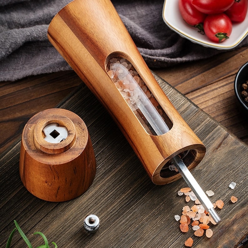 May include: A wooden salt grinder with a clear glass insert filled with pink Himalayan salt crystals. The grinder has a metal mechanism and is open, revealing the salt inside. The grinder is sitting on a wooden surface.