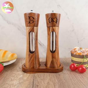 May include: A set of two wooden salt and pepper shakers with a clear glass insert. The shakers are engraved with the letters "D" and "K" and are sitting on a wooden base.