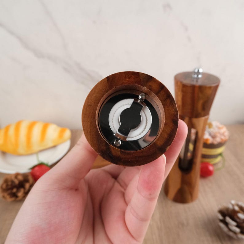 May include: Close-up of the bottom of a wooden salt or pepper grinder. The grinder has a clear plastic window to see the contents. The grinder has a metal grinding mechanism.