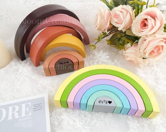 Könnte beinhalten: Holz-Regenbogen-Stapeltürme in Pastellfarben. Die Spielzeuge sind in einem Halbkreis angeordnet, wobei jede Schicht eine andere Farbe hat. Der Text "Rowyn" und ein Herz sind auf der untersten Schicht eines Regenbogens aufgedruckt. Der Text "dada" und ein Herz sind auf der untersten Schicht eines anderen Regenbogens aufgedruckt.