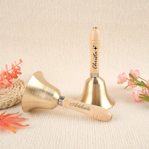 May include: Two brass-colored hand bells with wooden handles. One bell is upright, the other is lying down. The handle of the upright bell has the name "Christa" printed on it. The handle of the other bell has the name "Adelaide" printed on it.