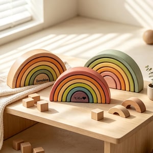 Puede incluir: Tres juguetes apilables de arcoíris de madera en tonos tierra suaves. Un conjunto lleva el nombre Zenobia, otro el nombre Willen con un símbolo de corazón. El set incluye varios cubos de madera pequeños y dos arcos de madera semicirculares.