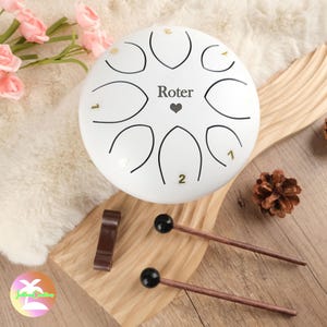May include: White handpan with the word "Roter" and a heart symbol in the center. The handpan has eight notes, numbered 1 and 2, and is resting on a wooden surface with two wooden mallets.