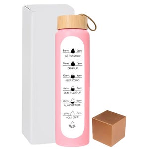Puede incluir: Una botella de agua rosa con tapa de bambú y un lazo para llevar. La botella tiene un horario impreso en blanco con frases y horarios motivacionales. Una caja blanca y un pequeño cubo color cobre también están en la imagen.
