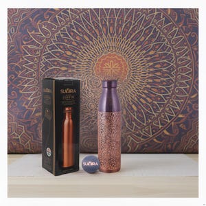 Peut inclure: Une gourde en cuivre avec un motif décoratif et un dessus violet. La bouteille est à côté de son emballage, d'un petit dessous de verre rond et d'un fond à motifs. L'emballage porte la mention "SUVIRA COPPER BOTTLE".