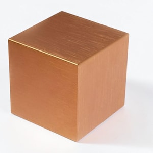 May include: A copper-coloured cube with a brushed metal finish. The geometric shape has sharp edges and a uniform colour, reflecting light across its surfaces. The cube is a simple, modern design.