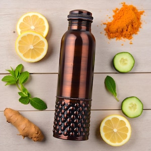 Puede incluir: Una botella de agua cilíndrica de cobre con un cuerpo superior liso y una sección inferior texturizada con hoyuelos y una banda de cuentas. La botella tiene una tapa de rosca a juego y un acabado metálico oscuro y pulido en posición vertical.
