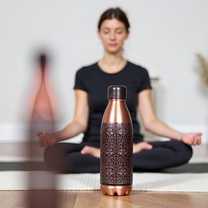 Puede incluir: Botella de agua de color cobre con un patr&oacute;n floral decorativo marr&oacute;n oscuro. La botella tiene una tapa y una base marrones. La botella mide aproximadamente 25 cm de alto y est&aacute; sobre una superficie de madera.