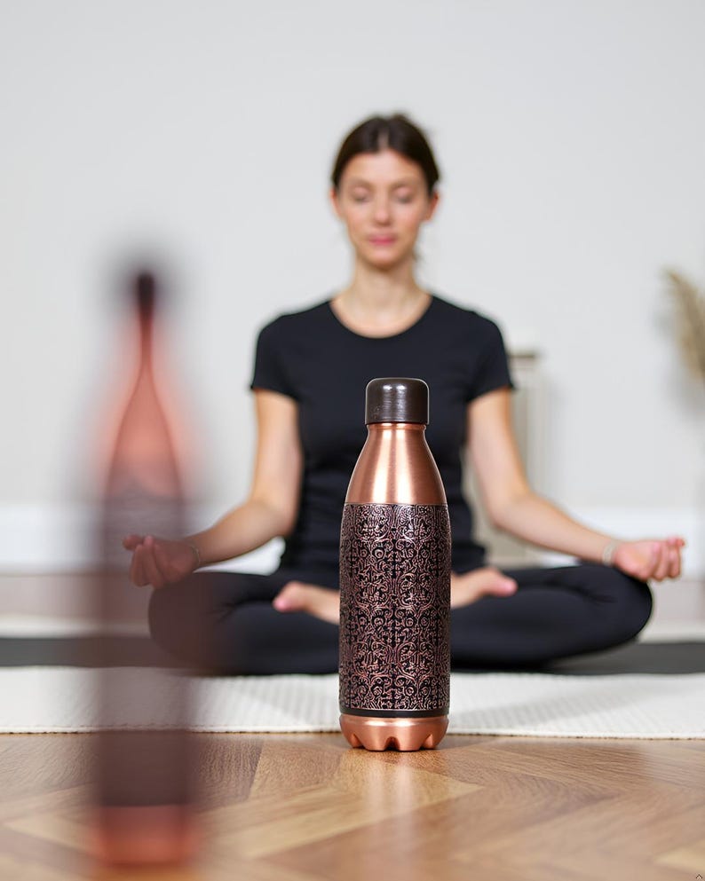 Puede incluir: Botella de agua de color cobre con un patr&oacute;n floral decorativo marr&oacute;n oscuro. La botella tiene una tapa y una base marrones. La botella mide aproximadamente 25 cm de alto y est&aacute; sobre una superficie de madera.
