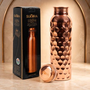 May include: A metallic copper water bottle with a textured honeycomb pattern on its exterior. The bottle features a narrow neck and a matching screw-top lid. Beside it stands a black rectangular box labelled Suvira Pure Copper Bottle.