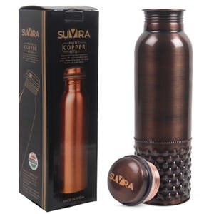 Puede incluir: Una botella de agua de cobre con tapa a juego y base decorativa. La botella est&aacute; junto a su caja negra original, que presenta la marca "SUVIRA" y las palabras "Pure Copper Bottle."