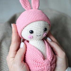 May include: A pink crocheted bunny doll with a white face and black eyes. The doll is wearing a pink crocheted hat and a pink crocheted blanket.