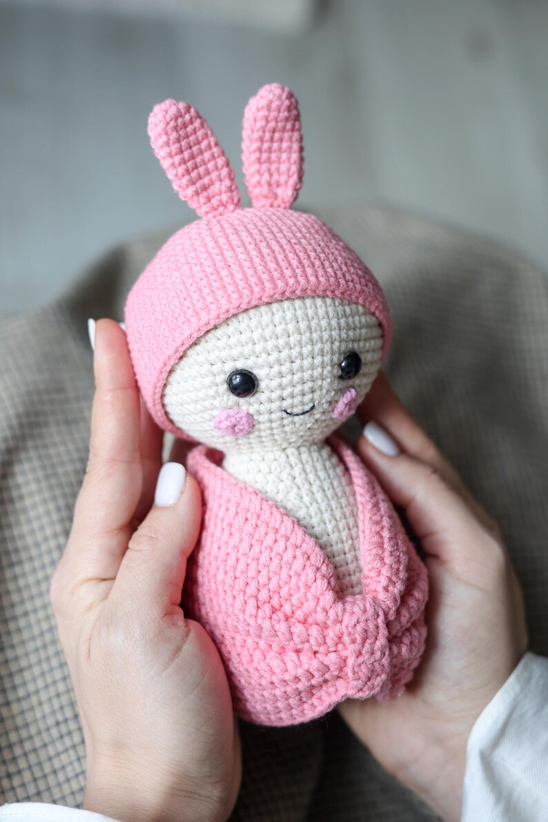 May include: A pink crocheted bunny doll with a white face and black eyes. The doll is wearing a pink crocheted hat and a pink crocheted blanket.
