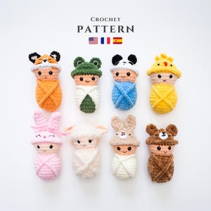 May include: Eight crocheted baby dolls in various animal costumes, including a fox, frog, panda, chicken, bunny, lamb, bear, and a plain doll. The dolls are all wrapped in blankets and have embroidered facial features. The dolls are arranged in two rows on a white background. The text "CROCHET PATTERN" is above the dolls, with the American, French, and Spanish flags.