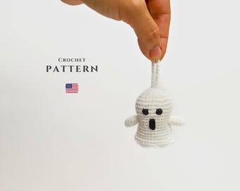 Puede incluir: Un adorno de fantasma blanco tejido a crochet con ojos negros y boca, colgando de una mano. El texto "CROCHET PATTERN" está sobre una pequeña bandera estadounidense.