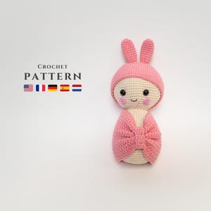 May include: Pink crocheted amigurumi doll with bunny ears and a bow. The doll has a smiling face with pink cheeks. The doll is wrapped in a pink blanket.