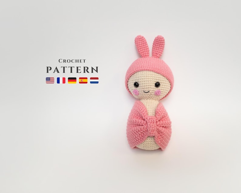May include: Pink crocheted amigurumi doll with bunny ears and a bow. The doll has a smiling face with pink cheeks. The doll is wrapped in a pink blanket.
