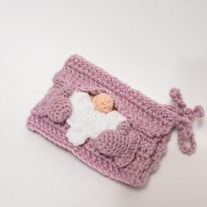 May include: A pink crocheted gnome with a white crocheted beard and a pink nose. The gnome is lying down with its arms crossed and its head resting on its arms.