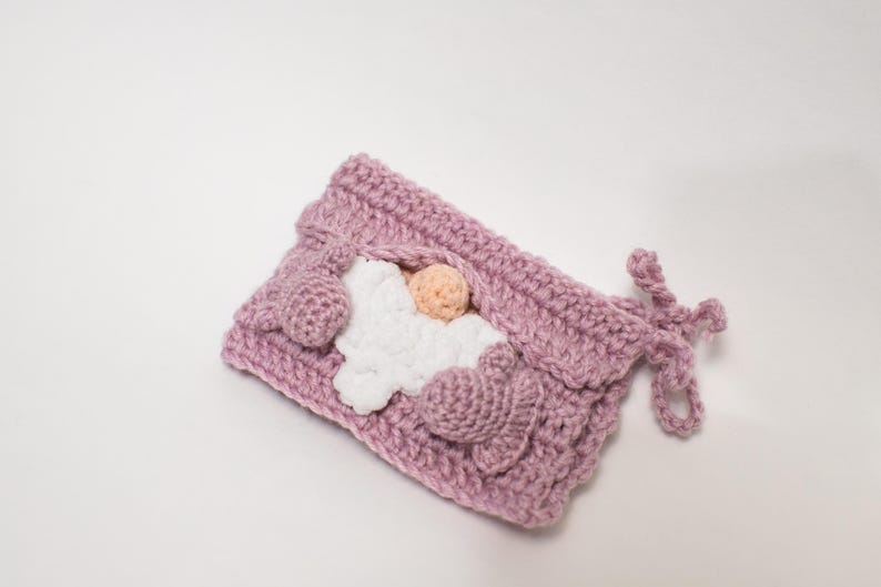 May include: A pink crocheted gnome with a white crocheted beard and a pink nose. The gnome is lying down with its arms crossed and its head resting on its arms.