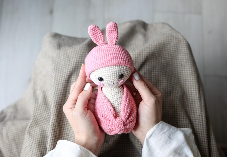 May include: A pink crocheted bunny doll with a white face and black eyes. The doll is wearing a pink crocheted hat with bunny ears.