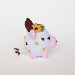 May include: A handmade crochet cow figurine. The white body has brown and yellow accents, with pink hooves and snout. It has brown horns and a sunflower crown. The cow has a small brown tail and black eyes.