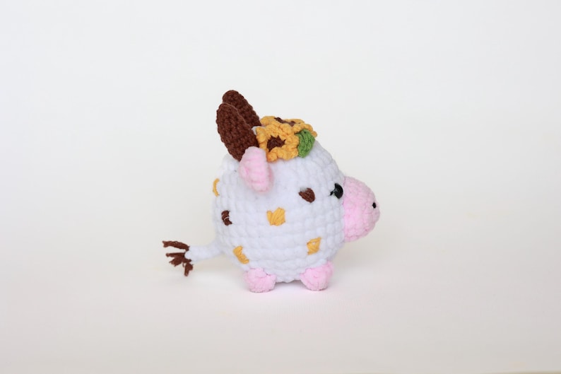 May include: A handmade crochet cow figurine. The white body has brown and yellow accents, with pink hooves and snout. It has brown horns and a sunflower crown. The cow has a small brown tail and black eyes.