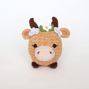 May include: A plush, light brown cow with dark brown horns and hooves. The cow has a white and green floral crown and black eyes. The soft, round toy is a handmade item, perfect for a child's room or nursery.