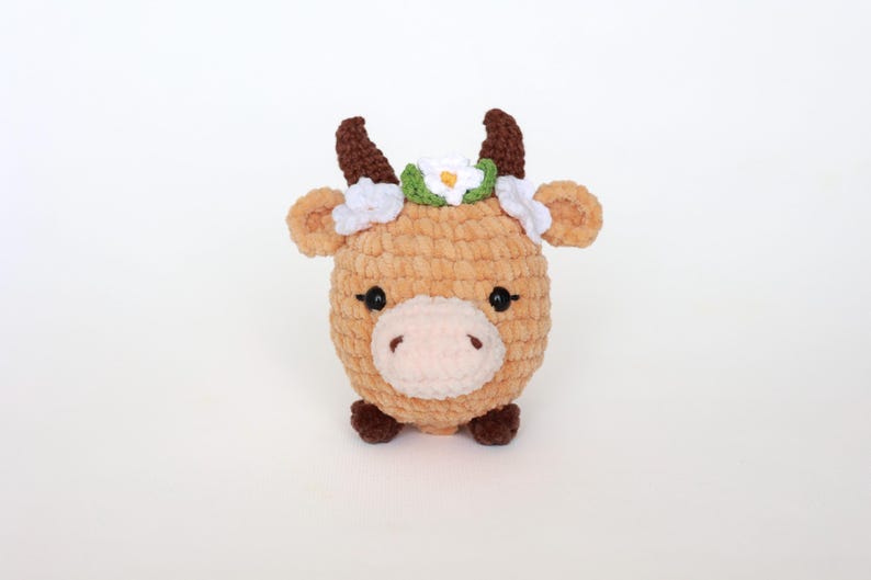 May include: A plush, light brown cow with dark brown horns and hooves. The cow has a white and green floral crown and black eyes. The soft, round toy is a handmade item, perfect for a child's room or nursery.
