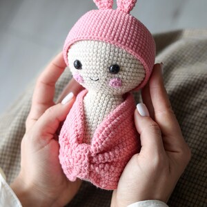 May include: A pink crocheted bunny doll wearing a pink hat and a pink blanket. The doll has a white face with black eyes and a pink nose.