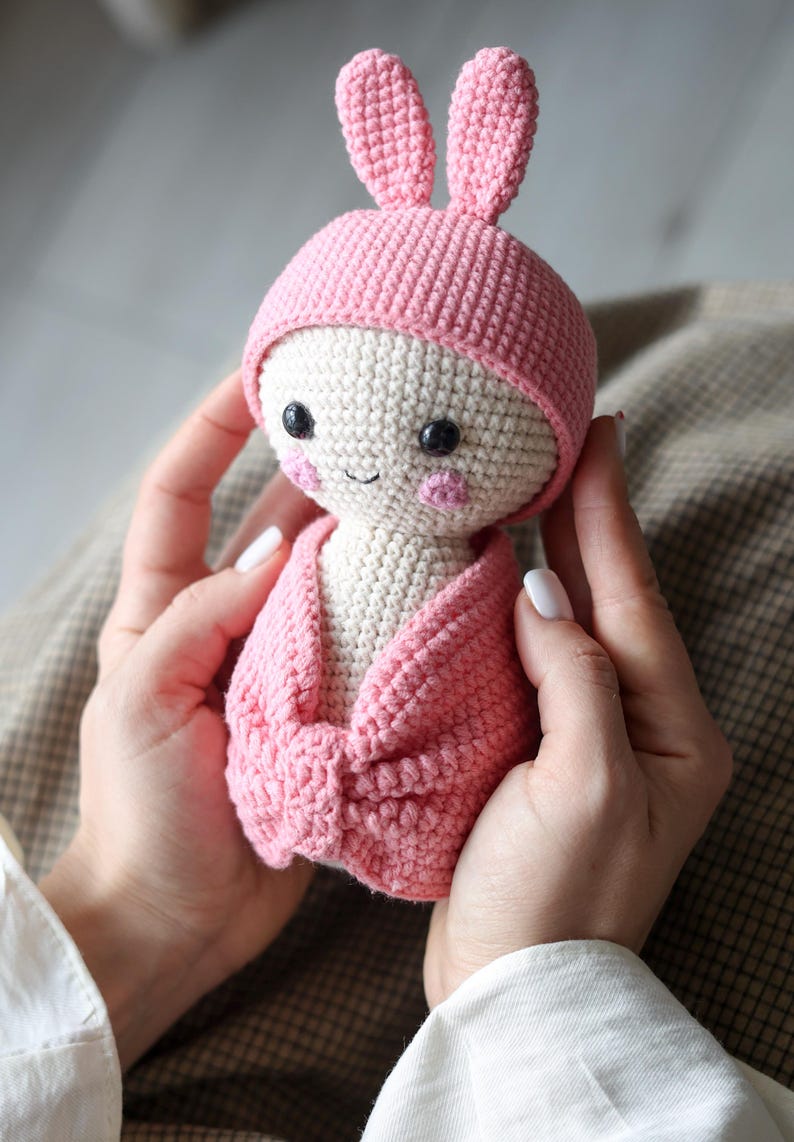 May include: A pink crocheted bunny doll wearing a pink hat and a pink blanket. The doll has a white face with black eyes and a pink nose.