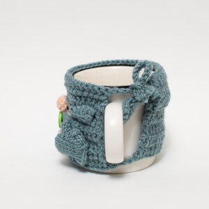 May include: A blue crocheted mug cozy with a flower and button embellishment. The cozy is on a white mug with a handle.