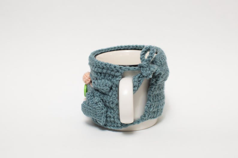 May include: A blue crocheted mug cozy with a flower and button embellishment. The cozy is on a white mug with a handle.