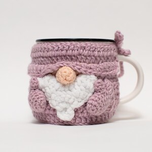May include: A pink crocheted mug cozy shaped like a gnome with a white beard and a pink nose. The cozy is on a white mug with a black rim.