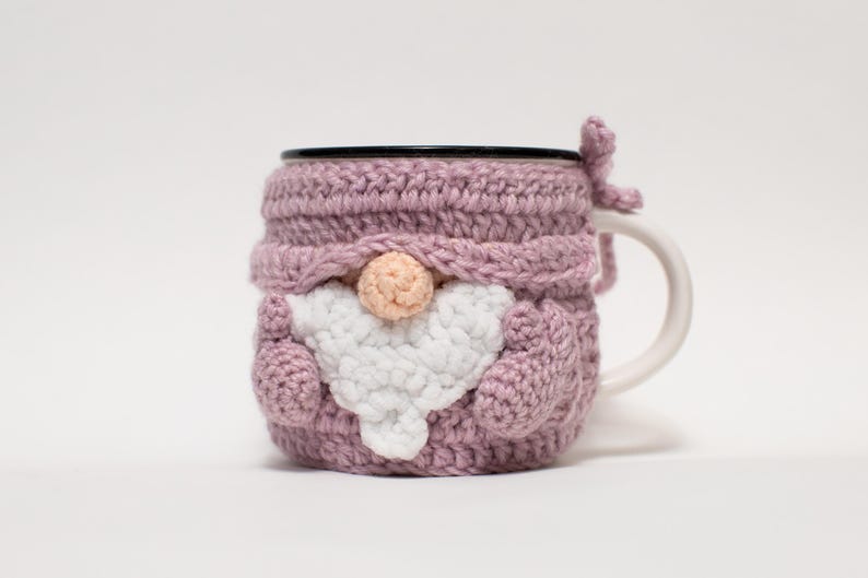 May include: A pink crocheted mug cozy shaped like a gnome with a white beard and a pink nose. The cozy is on a white mug with a black rim.
