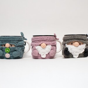 May include: Three crocheted mug cozies shaped like gnomes. The cozies are in blue, pink, and gray. Each gnome has a white beard and a button nose.