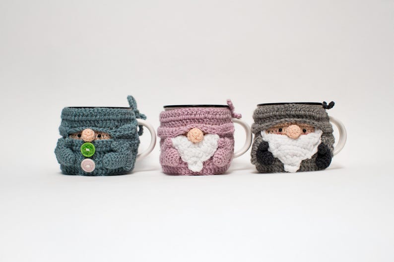 May include: Three crocheted mug cozies shaped like gnomes. The cozies are in blue, pink, and gray. Each gnome has a white beard and a button nose.