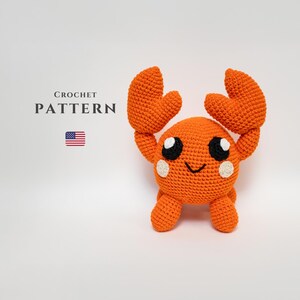 May include: An orange crochet crab with large claws and a smiling face. The crab has white accents around its eyes and on its cheeks. The image includes the text "CROCHET PATTERN" and a small American flag.