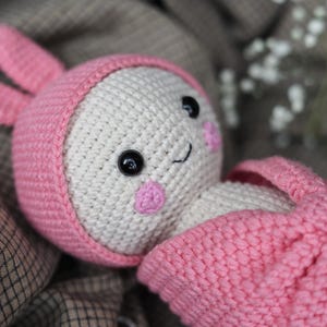 May include: A crocheted pink and white bunny toy with a pink hat and large black eyes. The bunny is lying on a brown and white checkered fabric.