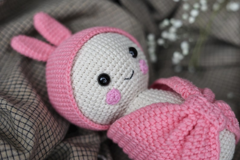 May include: A crocheted pink and white bunny toy with a pink hat and large black eyes. The bunny is lying on a brown and white checkered fabric.