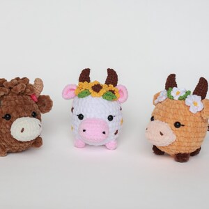 May include: Three handmade crochet cow plushies. The cows are brown, white, and tan, each with small horns and a white muzzle. The white cow has a sunflower crown, and the other two have flower crowns.