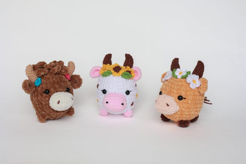 May include: Three handmade crochet cow plushies. The cows are brown, white, and tan, each with small horns and a white muzzle. The white cow has a sunflower crown, and the other two have flower crowns.