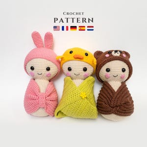 Puede incluir: Tres muñecas de bebé de crochet, cada una con un sombrero de animal diferente. Las muñecas están envueltas en mantas y tienen rasgos faciales bordados. Los sombreros son un conejito rosa, un pollito amarillo y un oso marrón.
