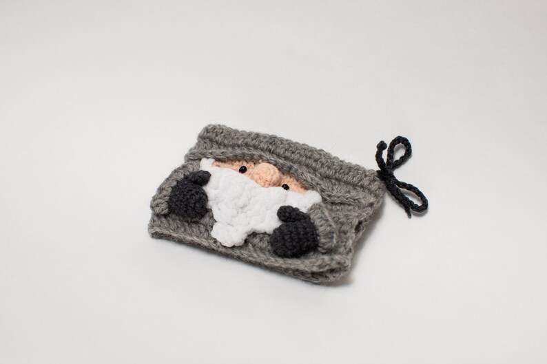 May include: Gray crocheted pouch with a black drawstring. The pouch features a crocheted face of a gnome with a white beard and black eyes.