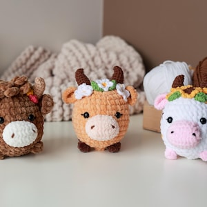 May include: Three handmade crochet cow plushies in brown, orange, and white. Each cow has a unique floral crown and a white muzzle. The plushies are on a white surface with yarn and a wooden box in the background.