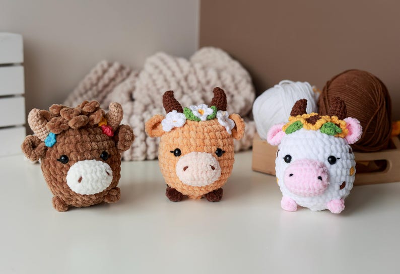 May include: Three handmade crochet cow plushies in brown, orange, and white. Each cow has a unique floral crown and a white muzzle. The plushies are on a white surface with yarn and a wooden box in the background.
